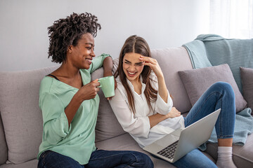 Cheerful young woman using laptop and looking at happy friend while sitting at sofa and discussing homework assignment in cozy room. Two excited women shopping online