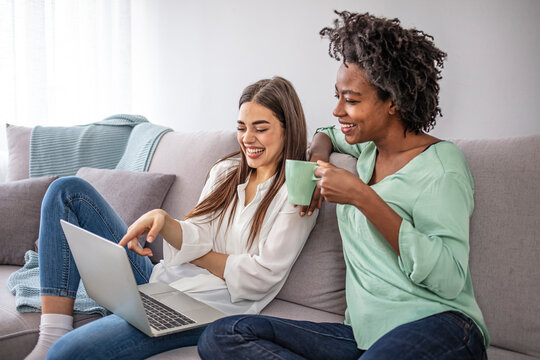 Two Diverse Friends Relaxing On Comfortable Couch And Watching Film Online On Laptop Device Using Home Wireless Internet.Multicultural Young Women Working Together On Common Blog At Computer