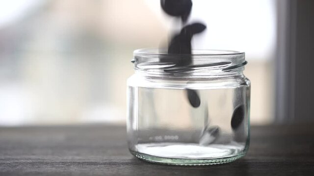 Ukrainian coins falling into the glass jar on wooden table near window, close up. Slow motion, a pile of silver coins falls into an empty glass jar on a window background. Ukraine