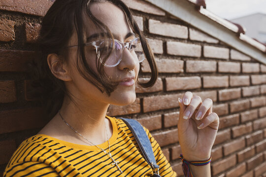 Teenager girl wearing colorful clothes and transparent clear eyeglasses in front of a red brown brick wall posing playing with her hair. Downtown city centre autumn concept.