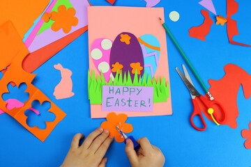 child hands makes a card, application, Easter bunny and eggs from colored paper. Top view