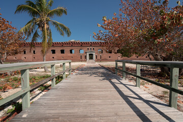 Gate of an old military fort with a wooden walk in the foreground. Big brick walls and green palm...