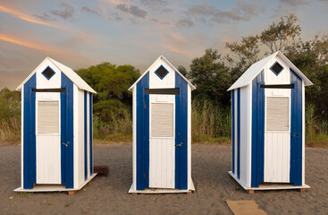 Fototapeta na wymiar Three pretty wooden toilet houses stand on a beach near Marbella in southern Spain. It's an early summer morning at sunrise. There are orange clouds in the sky.