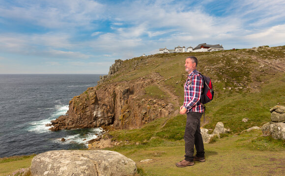 A backpacker hiker stands on the high cliffs on the Atlantic coast of Cornwall at Land's End. He looks at the sea. In the background are the buildings for the tourists.