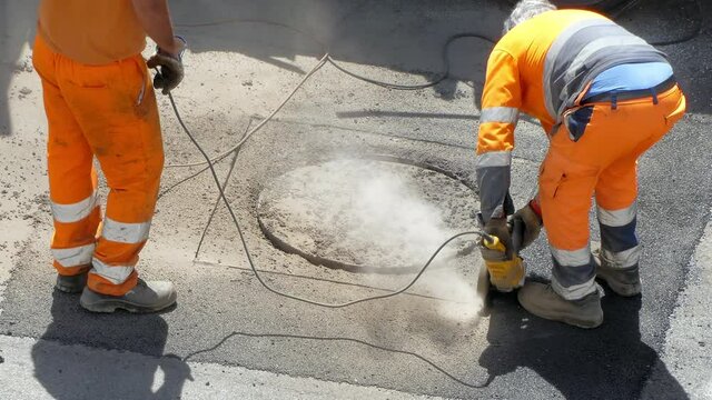 Drilling works on the road, workmen in orange uniform working on construction site on the sidewalk. Male professional workers using powerful cutting machine for breaking old layer of cement and