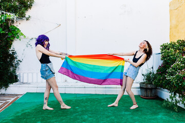 Naklejka premium Two women with rainbow flag on a backyard. Couple Young lesbian girls holding a rainbow flag and hugging happily. A pair of lesbian women