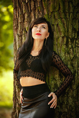 sexy brunette girl in a lace blouse and leather skirt with high heels posing near a tree in the...