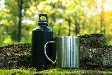 A metal flask and a mug stand on an old tree stump in the forest. Eco friendly, camping and healthy lifestile concept.