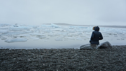 Person sitting on a rock by a lake with ice and surrounded by fog in winter. Jokulsarlon Glacial Lake