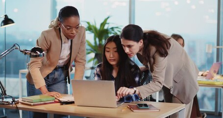 Three multi-cultural young women discussing computer project on laptop negotiating business plans coworking in corporate modern office.
