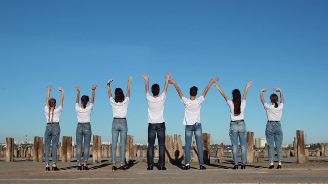 Seven people in white T-shirts and jeans stand with their backs to the camera and waving their hands, standing on the seashore against the blue sky. Shooting at 205 fps.