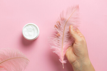 Creative composition with woman’s hand holding pink ostrich feather and protective hand cream in round jar on pink pastel background. Skin cate concept.