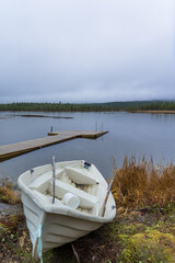 Old, dirty, used rowing boat laying next to a small finish lake in Inari, Finland, Lapland. Wooden pier at a relaxing, tranquil peaceful rainy day at a Cottage with green forrest-upright