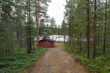 Path leads to beautiful red wooden cottage next to a stunning lake. Remote lonely cabin in Lapland, Finland in fresh green pine forest. Self sufficient hut for relaxing vacation in Inari