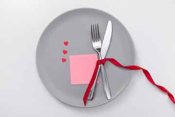Valentine's day festive table setting, flat lay with red heart shape on gray plate, fork, knife and red ribbons on white table. Love dating concept. Place your text and copy space. High quality photo