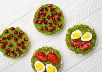 Rice cakes with avocado mash and eggs with tomato and pomegranate seeds on white wooden background