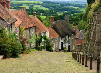 Fototapeta na wymiar View To Old Limestone Houses On A Cobble Street At Gold Hill In Shaftesbury England With A Beautiful Rural Landscape In The Background On An Overcast Summer Day