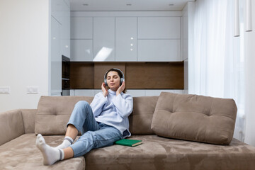 Young beautiful woman is resting at home, sitting on the couch, and listening to music on headphones