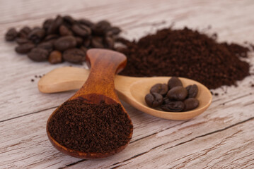 Dark and light colored spoons contrasted one filled with grounded coffee beans and one with coffee beans, a pile of coffee beans and grounded beans in the background