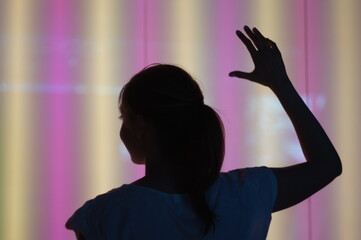 A young woman is raising her hand looking to the left. We see her from back and don't see her face. The background is colourful neon wall.