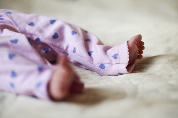 Lifestyle image of cute adorable baby girl (daughter) feet with tiny toes on bed close up. Selective focus. Legs are in pink pants with purple heart. Copy space for message. 