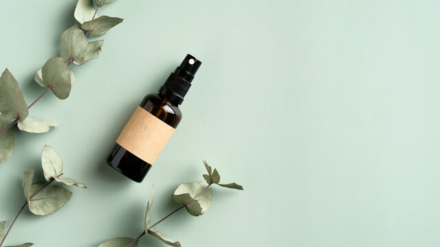 Amber glass spray cosmetic bottle and eucalyptus leaves on green background. SPA herbal beauty product packaging design, branding.
