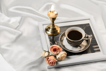 Coffee in bed, romantic atmosphere. Flowers, a burning candle, and a cup of coffee on white on a silk bed. Home cozy interior, lifestyle. Selective focus,copy space.