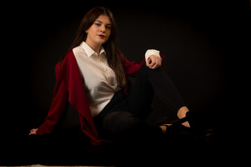 Mujer guapa y atractiva posa sentada sobre fondo oscuro vestida con camisa blanca chaqueta roja y...