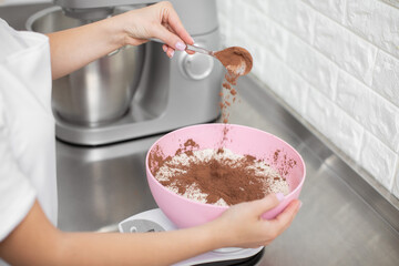Close up cropped shot of hands of professional woman chef, pouring cacao powder into the bowl with white flour. Making macarons, cakes and desserts