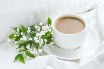 Obraz na płótnie Canvas Cup of coffee on the bed with white flowers and green leaves. White bed linen, good morning concept.