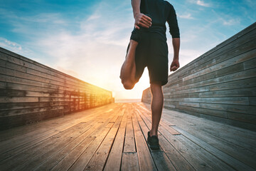 Young slim sporty man warming and stretching legs before run outdoors at sunset or sunrise. Athletic man in black sportswear doing fitness stretching exercises. Sport and healthy lifestyle.