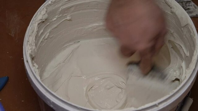 Stir the gray putty with the White Bucket. Human hand. Iron spatula. Artificial light. Preparation of the base. Internal construction and finishing works. Home repairs. Close-up.