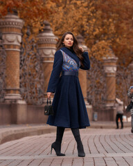 Fototapeta na wymiar Elegant woman wearing blue coat and walking city street on autumn or fall day against town park fence. Pretty girl with makeup and wavy brunette hair