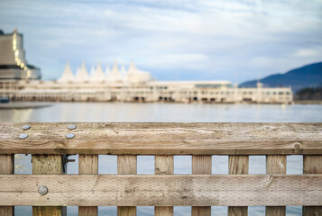 Fototapeta premium Weathered wood railing with blurry waterfront buildings and ocean background and cloudy sky. Defocused and abstract Vancouver Harbour building or Canada Place. Selective focus.