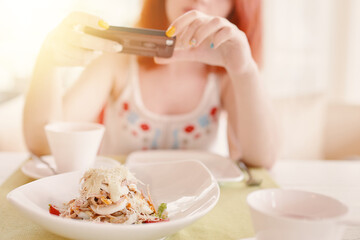 Woman takes photos of salad on smartphone camera. Red-haired girl in cafeteria at table with white ceramic dishes. Salad of eggs cheese and other ingredients. Summer lunch on terrace.
