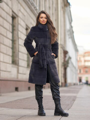 Fototapeta premium Elegant stylish woman with long brunette wavy hair wearing black trousers, pullover, shoes and fur coat walking city street on a sunny day