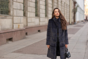 Fototapeta premium Elegant stylish woman with long brunette wavy hair wearing black trousers, pullover, shoes and fur coat walking city street on a sunny day