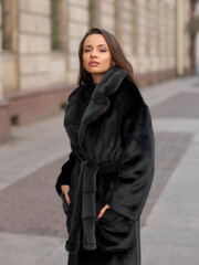 Fototapeta premium Elegant stylish woman with long brunette wavy hair wearing black trousers, pullover, shoes and fur coat walking city street on a sunny day