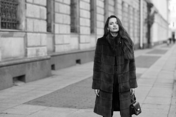 Elegant stylish woman with long brunette wavy hair wearing black trousers, pullover, shoes and fur coat walking city street on a sunny day