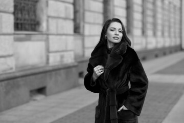 Elegant stylish woman with long brunette wavy hair wearing black trousers, pullover, shoes and fur coat walking city street on a sunny day