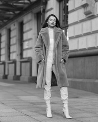 Elegant stylish woman with long brunette wavy hair wearing white trousers, pullover and shoes and purple fur coat walking city street on a sunny day