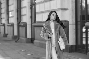 Obraz premium Elegant stylish woman with long brunette wavy hair wearing white trousers, pullover and shoes and purple fur coat walking city street on a sunny day