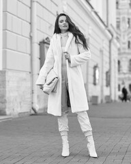 Elegant stylish woman with long brunette wavy hair wearing white trousers, pullover and shoes and white fur coat walking city street on a sunny day