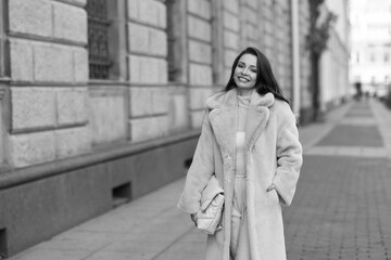 Naklejka premium Elegant stylish woman with long brunette wavy hair wearing white trousers, pullover and shoes and pink fur coat walking city street on a sunny day