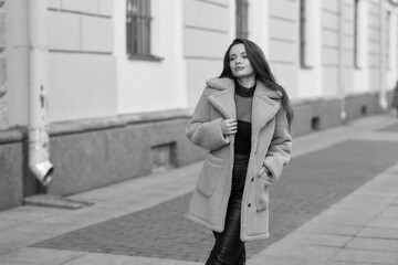 Obraz premium Elegant stylish woman with long brunette wavy hair wearing black trousers, pullover, shoes and pink fur coat walking city street on a sunny day