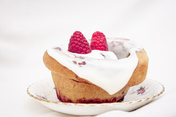 Exquisite cinnamon roll with white glaze and red fruits on top on a white porcelain plate on a white table.