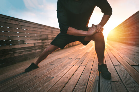 Sporty man stretching leg adductor muscles and warming up for training outdoors at sunset or sunrise. Athletic man in black sportswear doing fitness stretching exercises. Sport and healthy lifestyle
