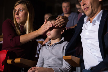 Young woman looking shocked while watching movie in cinema, closing eyes of her preteen son with her hand. Concept of parental guidance and protection