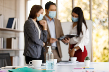 coronavirus prevention in the office. Bottle of alcohol gel on a table with a soft-focus background of a group of Asian businesspeople wearing face masks standing reading working discussion
