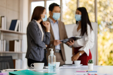 coronavirus prevention in the office. Bottle of alcohol gel on a table with a soft-focus background of a group of Asian businesspeople wearing face masks standing reading working discussion